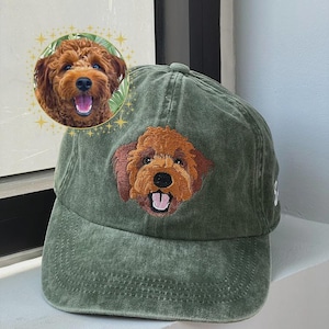 May include: An olive green baseball cap featuring an embroidered patch of a brown curly-haired dog face with a black nose and an open mouth showing a pink tongue. The cap has a distressed fabric texture and a structured six-panel design.