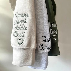 May include: Close-up of three sweatshirt sleeves in cream, gray, and olive green. Each sleeve features embroidered names in cursive script. The cream sleeve has names Danny, Jacob, Addie, and Rhett, with a green heart. The gray sleeve has Theo and Bowen. The olive sleeve has Madeline and Henry, with a white heart.