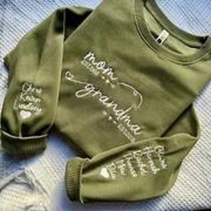 May include: An olive green sweatshirt with white embroidered text. The text on the front reads "mom grandma" with established dates. The left sleeve has the names "Chris Kevin Lindsey" embroidered. The right sleeve has the names "Chris Hannah Rachel Thomas Ella" embroidered.
