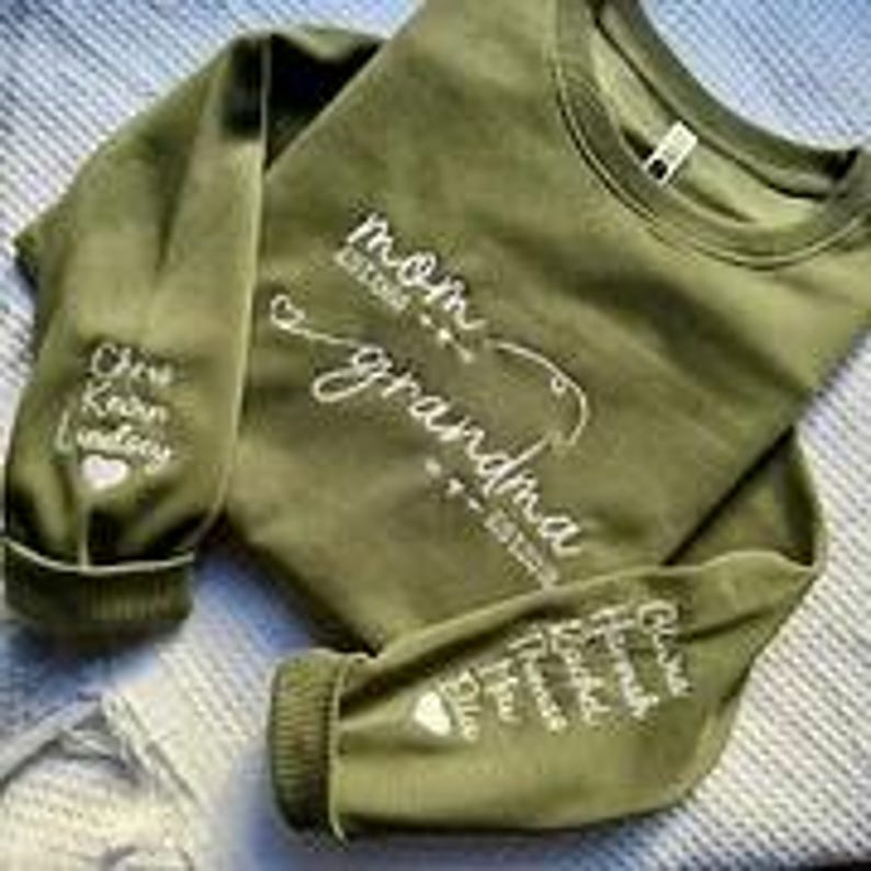 May include: An olive green sweatshirt with white embroidered text. The text on the front reads "mom grandma" with established dates. The left sleeve has the names "Chris Kevin Lindsey" embroidered. The right sleeve has the names "Chris Hannah Rachel Thomas Ella" embroidered.