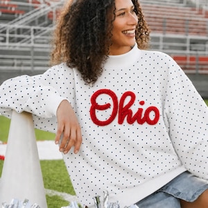 Könnte beinhalten: Ein weißes Langarm-Sweatshirt mit schwarzem Punktemuster und Stehkragen. Auf der Vorderseite befindet sich das Wort Ohio in großen, strukturierten roten Chenille-Buchstaben. Das Kleidungsstück hat eine lockere Passform mit gerippten Bündchen.