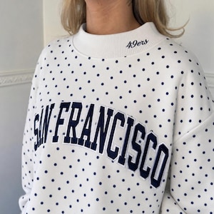 May include: A white sweatshirt featuring a navy blue polka dot pattern throughout. The front displays the text SAN FRANCISCO in large navy blue embroidered letters, while the ribbed mock neck collar includes the embroidered text 49ers.