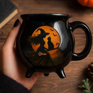 May include: Black cauldron-shaped mug with a handle and three small feet. The mug features a Halloween design with a witch on a broomstick, a black cat, and the name "Olivia" in orange against an orange moon background.