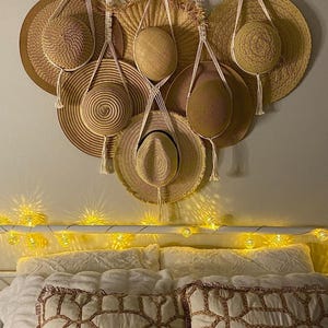May include: A wall hanging with seven straw hats hanging from macrame cords. The hats are various sizes and shapes. The wall hanging is decorated with a string of white lights.
