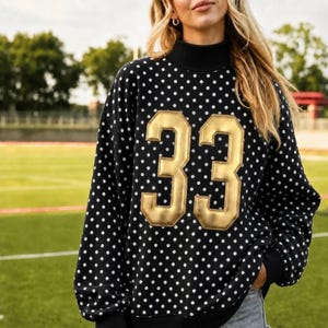 Könnte beinhalten: Ein schwarzes Langarm-Sweatshirt mit weißem Punktemuster und einer großen, goldfarbenen Zahl 33 auf der Vorderseite. Das Kleidungsstück verfügt über einen hohen Stehkragen und gerippte Bündchen, was für eine strukturierte Passform sorgt.