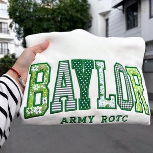 May include: A white sweatshirt featuring the word BAYLOR in large, green-bordered applique letters with various patterns including floral, stripes, and polka dots. Below the main text, the words ARMY ROTC are embroidered in green thread.
