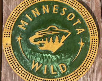 May include: A circular wooden cribbage board featuring the Minnesota Wild logo in the center. The board has a natural wood outer ring with drilled holes for scoring, marked with Start, Finish, and SS. The center is filled with green resin.