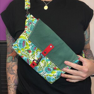 May include: A green and floral patterned crossbody bag with a red strap and a flap closure. The bag has a smaller green pocket on the front.