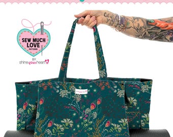 May include: A teal Yoga Tote Grande sewing pattern with a floral design. The tote bag has a black yoga mat inside. The text "Yoga Tote Grande Sewing Pattern" is at the top of the image. The bag is held by a person.