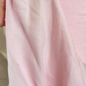 May include: Close-up of a soft, light pink fabric. The fabric is slightly wrinkled, with visible texture and a subtle sheen. The material appears to be lightweight and suitable for clothing or home decor projects.