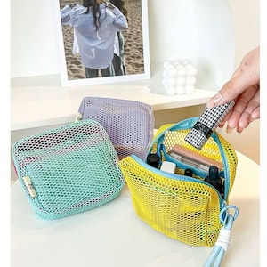 May include: Three mesh cosmetic bags in teal, lavender, and yellow. The yellow bag is open, revealing makeup products. A hand holds a lipstick with a black and white pattern. The bags are on a white surface.