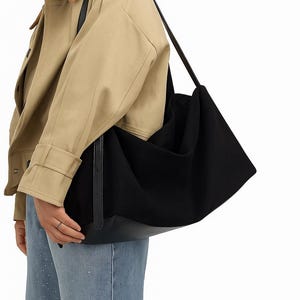 May include: A large black tote bag with a leather bottom and long straps. The bag is carried over the shoulder. The person in the image is wearing a tan jacket and blue jeans.
