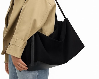 May include: A large black tote bag with a leather bottom and long straps. The bag is carried over the shoulder. The person in the image is wearing a tan jacket and blue jeans.