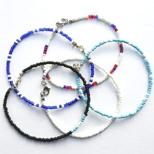 Peut inclure: Plusieurs bracelets de cheville perlés de différentes couleurs. Les bracelets sont ornés de petites perles blanches, noires, bleues et rouges, avec des fermoirs argentés. Les bracelets sont disposés en cercle sur un fond blanc.