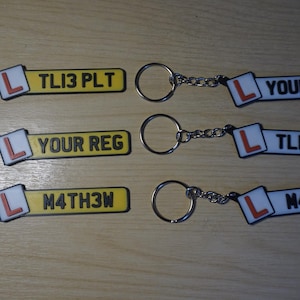 May include: Keychains with license plate designs. Each keychain has a silver chain and ring. The license plates are yellow with black text and a white square with a red "L" symbol. The text on the plates reads "TL13 PLT", "YOUR REG", and "M4TH3M".