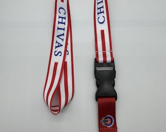 Op de afbeelding: Een rood-wit gestreepte lanyard met het woord "CHIVAS" in blauw gedrukt. De lanyard heeft een zwarte plastic sluiting en een metalen clip. Een klein rood gedeelte heeft een logo.