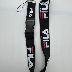 May include: Black lanyard with the word "FILA" in white and red lettering. The lanyard has a black plastic buckle and a silver-colored metal clasp. The lanyard is designed to be worn around the neck.