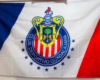 Op de afbeelding: Een witte vlag met een blauw en rood diagonaal ontwerp. Het midden toont een cirkelvormig embleem met een ridderhelm, een schild en de woorden "CLUB DEPORTIVO GUADALAJARA" in het blauw.