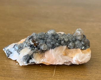 May include: A cluster of small, gray, and brown crystals on a bed of pink and white barite. The crystals are clustered together in a dense formation, creating a visually interesting and textured piece.