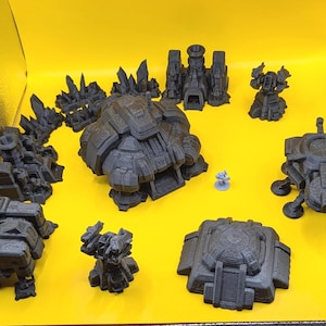 May include: A collection of dark gray 3D-printed miniature structures, including buildings, vehicles, and terrain elements. The pieces are arranged on a bright yellow surface, showcasing various shapes and sizes, ideal for tabletop gaming or model building.