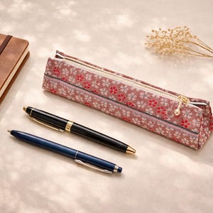以下が含まれることがあります： 茶色の革張りのノート、花柄のペンケース、2本のペンが明るい色の表面に配置されています。1本のペンは黒と金色のアクセント、もう1本は青色です。ペンケースはピンクの花柄です。