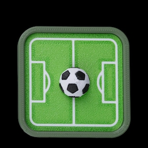 May include: A miniature soccer field with a soccer ball in the center. The field is green with white lines and a dark green border. The soccer ball is black and white.