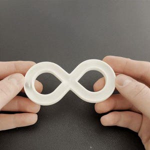 Pode incluir: Um símbolo de infinito branco, segurado entre duas mãos, contra um fundo escuro. O símbolo é um loop contínuo, representando a eternidade ou possibilidades infinitas. O objeto é feito de um material sólido e opaco.