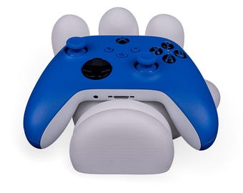 Könnte beinhalten: Ein blau-weißer Videospiel-Controller, der auf einem weißen, pfotenförmigen Ständer ruht. Der Controller hat schwarze Knöpfe und einen schwarzen Joystick. Der Ständer hat ein glattes, abgerundetes Design.