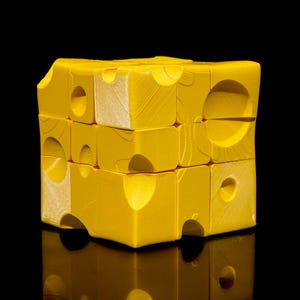 Puede incluir: Un cubo de rompecabezas amarillo con un diseño similar al queso, con agujeros circulares e irregulares. El rompecabezas está sobre un fondo negro, con un reflejo visible debajo. Un juguete divertido y único.