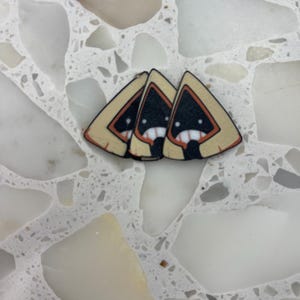 Peut inclure: Trois épinglettes triangulaires présentant un graphique stylisé d'une silhouette sombre à capuche avec une bordure beige, un intérieur noir, deux yeux blancs et une bouche blanche avec des dents. Les épinglettes sont alignées en rangée.
