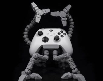 May include: A white Xbox controller with a gray, articulated stand. The stand has four legs and two arms that extend from the top of the controller. The arms are positioned in a way that suggests the controller is holding a weapon.