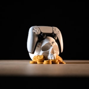 Puede incluir: Una escultura de cráneo humano blanco que descansa sobre un montón de monedas circulares de color dorado, con un mando de videojuego blanco y negro equilibrado sobre el cráneo. El objeto está situado sobre un fondo negro sólido y oscuro.