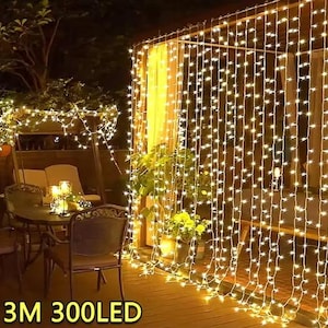 May include: Outdoor scene illuminated by warm white LED string lights. The lights are arranged in a curtain-like pattern, creating a cozy ambiance. The text "3M 300LED" is visible at the bottom. The scene includes a table, chairs, and plants.