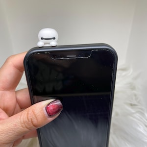 May include: A black smartphone with a white Baymax figure attached to the top. The phone is held in a hand with red and black nail polish. The background is a soft, white, fluffy material.