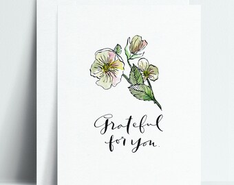 May include: A white greeting card with a watercolor illustration of delicate white and yellow flowers with green leaves. The card has the handwritten text "Grateful for you."