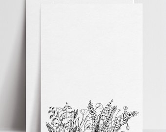 Puede incluir: Juego de papelería blanco con una tarjeta en blanco y un sobre. La parte inferior de la tarjeta está decorada con un dibujo lineal negro de varias flores silvestres y follaje. El papel tiene un acabado texturizado.