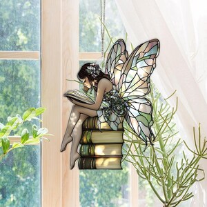 May include: A decorative fairy suncatcher with a stained-glass effect. The fairy is depicted reading a book while sitting on a stack of books. The fairy has large, colorful wings and is suspended by a chain. The color palette includes greens, browns, and pastels.