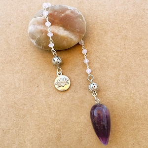 May include: A silver-toned chain features small pale pink beads, a round metal charm with an engraved lotus flower, and a teardrop-shaped deep purple amethyst crystal pendant. The chain is draped over a smooth, rounded, tan-colored stone.