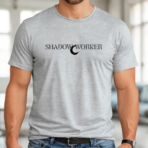May include: A light gray t-shirt with the text "SHADOW WORKER" in black, with a crescent moon graphic. The shirt is made of a soft material and has short sleeves. The model is wearing blue jeans and a black watch.