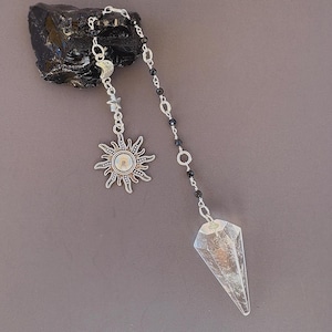 Peut inclure: Un pendule en cristal transparent à facettes fixé à une chaîne argentée avec de petites perles noires, une breloque étoile, une breloque croissant de lune et une breloque soleil avec une incrustation irisée, ancré par une pierre noire.