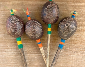 May include: Four maracas with dark brown, oval-shaped, hollowed-out gourd bodies featuring etched floral patterns. Each handle is made of wood and wrapped with vibrant orange, yellow, blue, and green thread, topped with small, colorful feather accents.