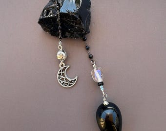 May include: A black obsidian stone piece attached to a silver-toned chain featuring a crescent moon and star charm, a small iridescent bead, and a smooth, teardrop-shaped black stone pendant hanging at the end of the chain assembly.