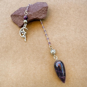 May include: A silver-toned chain pendulum featuring a teardrop-shaped purple amethyst stone, a decorative silver bead, and a small silver charm of a fairy sitting on a crescent moon. The chain is accented with small purple beads throughout its length.