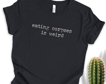Op de afbeelding: Zwart T-shirt met de tekst "eating corpses is weird" in een witte, typemachine-achtige letter. Het shirt is gemaakt van een zacht materiaal. Een groene cactus in een witte pot staat op de achtergrond.
