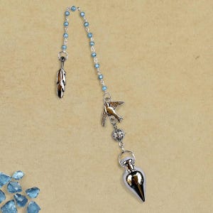 May include: A silver-toned pendulum with a bird and feather charm, connected by a chain of blue beads. The pendulum has a teardrop-shaped weight. The background is a light tan color.