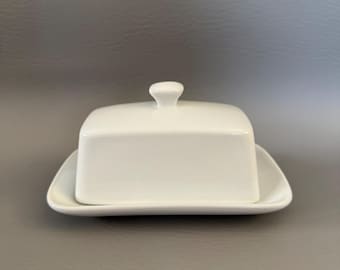 May include: A white ceramic butter dish with a lid and a small handle on top. The dish is rectangular with rounded edges and sits on a matching tray. The design is simple and elegant, suitable for kitchen or dining use.