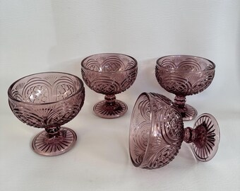May include: Four vintage amethyst-colored glass dessert bowls. Each bowl features a detailed, embossed pattern on the bowl and stem. The bowls are arranged on a white surface, with one bowl tipped on its side.