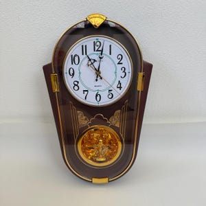 May include: A wall clock with a dark brown frame and gold accents. The white clock face features black Arabic numerals and the text "RHYTHM" and "QUARTZ". Below the dial, a gold-toned pendulum mechanism is visible behind a clear glass panel.