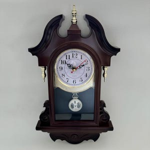 May include: A dark brown wooden wall clock with a decorative design. The clock face is white with black numerals and gold-colored hands. The clock has a pendulum and a shelf at the bottom.