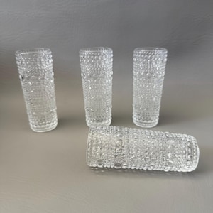 May include: Four clear glass tumblers featuring a textured surface with a repeating pattern of raised circular dots and small bubbles. Three of the tumblers are standing upright in a row, while one tumbler is positioned horizontally in front.